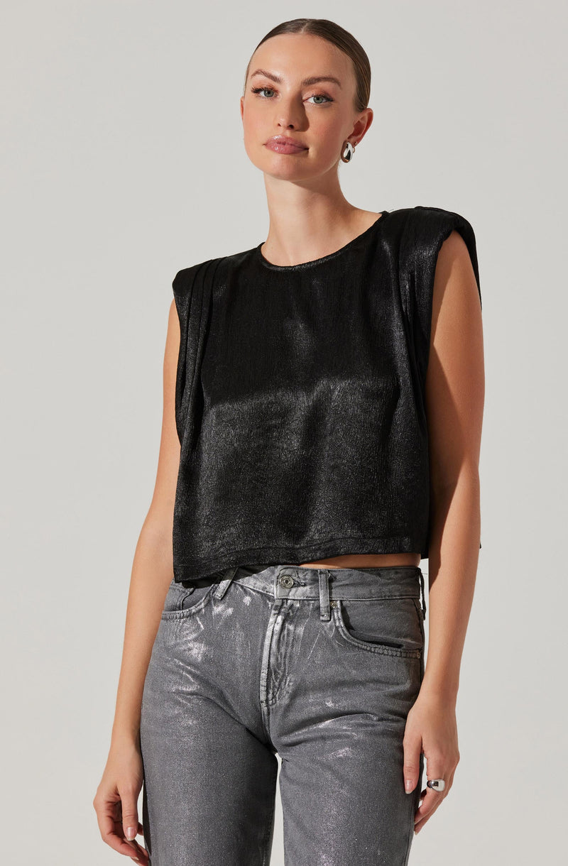 A woman is wearing a sleeveless black cropped top with a shiny, leather-like texture and slightly padded shoulders. She pairs it with high-waisted, metallic silver jeans.