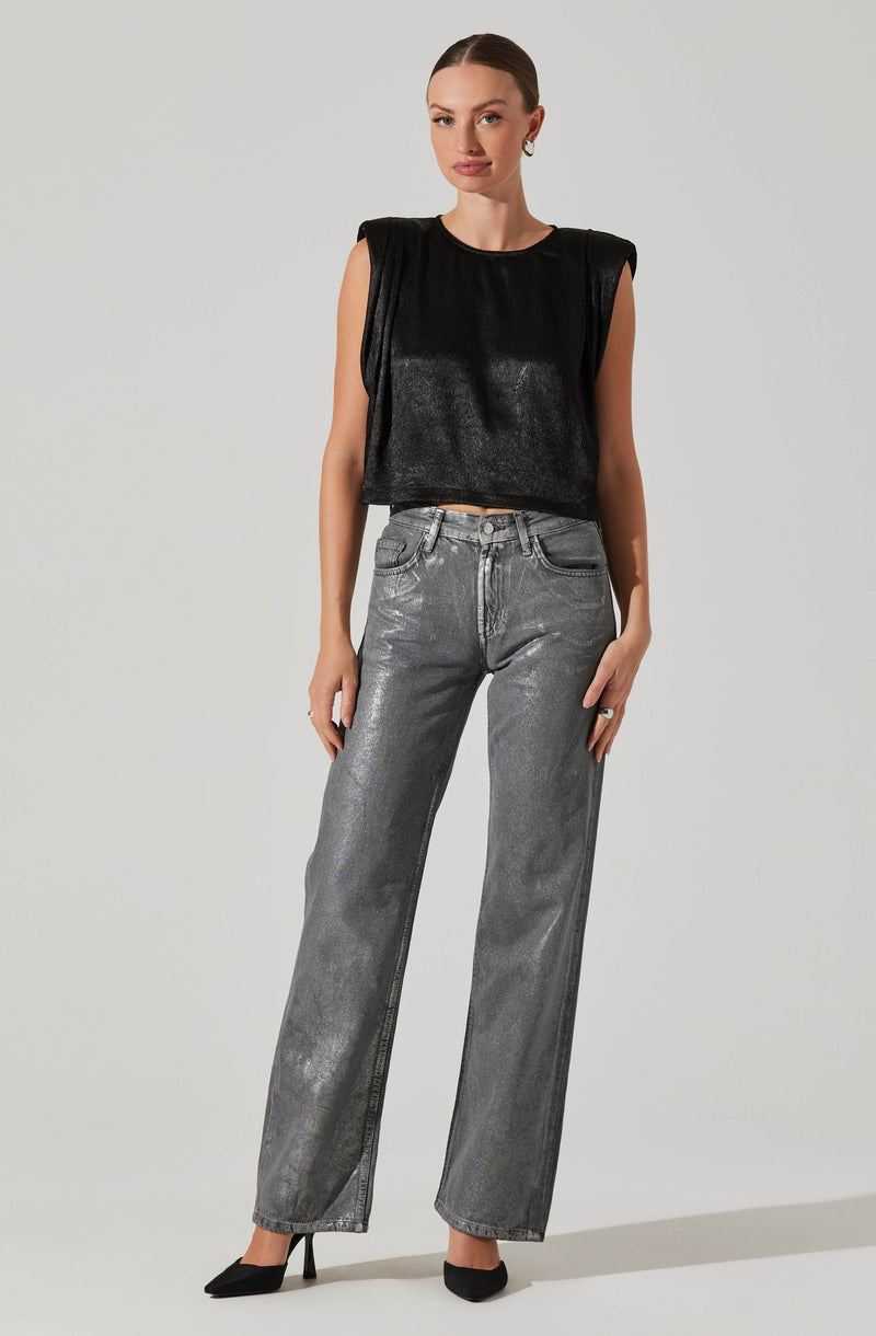 A woman is wearing a sleeveless black top with broad shoulder pads and shiny fabric, paired with high-waisted, straight-leg gray jeans that have a slightly metallic sheen. She completes her outfit with black pointed-toe high heels.
