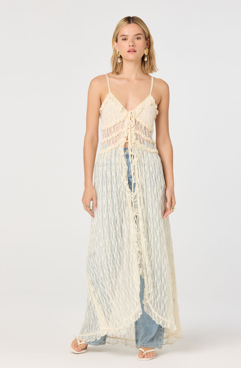 A woman is wearing a cream-colored, sheer, sleeveless top with thin spaghetti straps, featuring lace detailing and a ruffle trim along the neckline and hem. She pairs it with high-waisted, wide-leg jeans that are visible beneath the see-through fabric of her top. She also has on large gold hoop earrings and rings on her fingers.