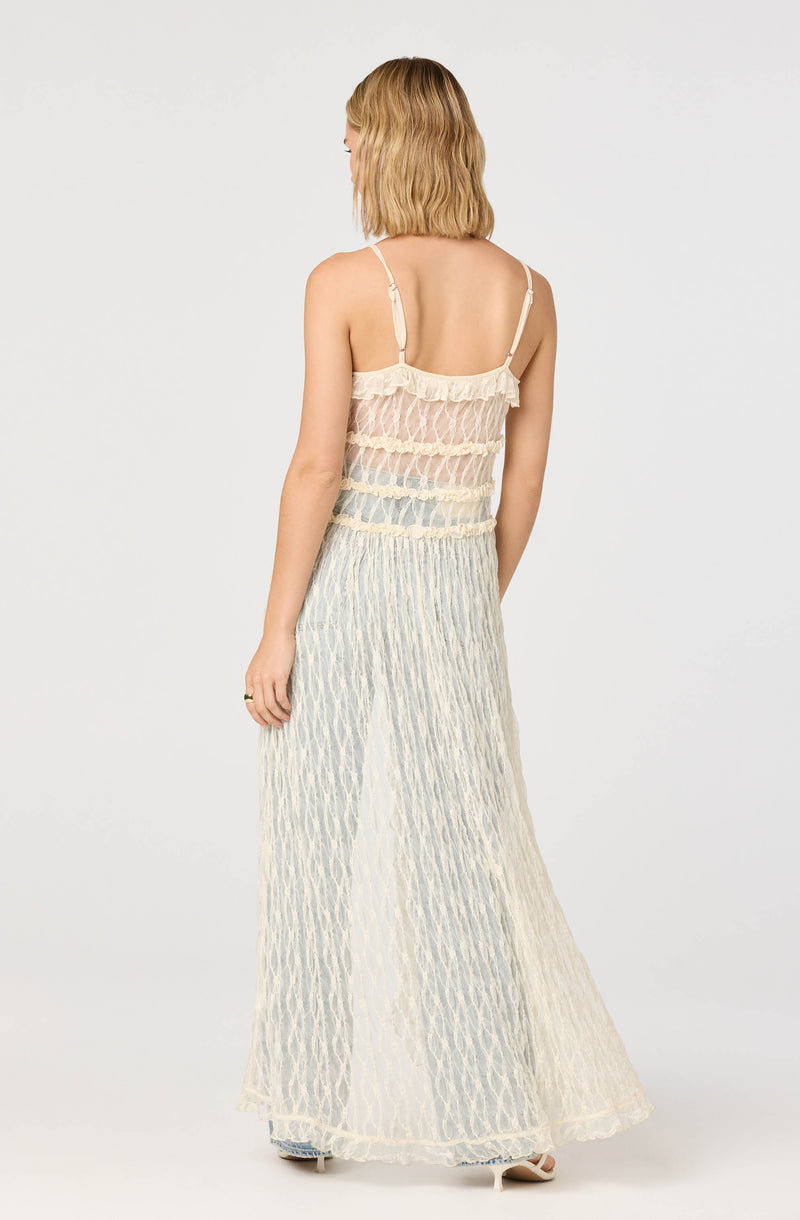 A woman is wearing a sheer, sleeveless, cream-colored top with thin spaghetti straps and a ruffled neckline. She is also wearing wide-leg, high-waisted pants made of a lightweight, semi-transparent fabric with a vertical striped pattern.