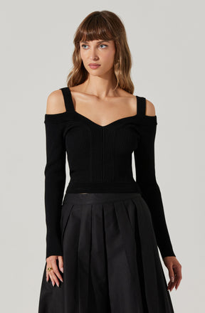 A woman is wearing an off-the-shoulder black ribbed sweater with wide straps and a fitted bodice. She pairs it with a high-waisted, pleated black skirt.