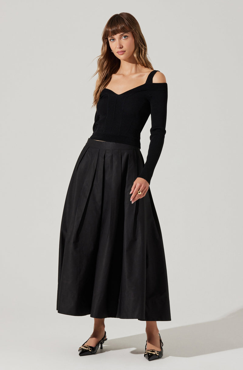 A woman is wearing a black off-the-shoulder sweater with long sleeves and a fitted design. She pairs it with a high-waisted, full-length black skirt featuring pleats. She completes the outfit with black pointed-toe heels decorated with a metallic detail.