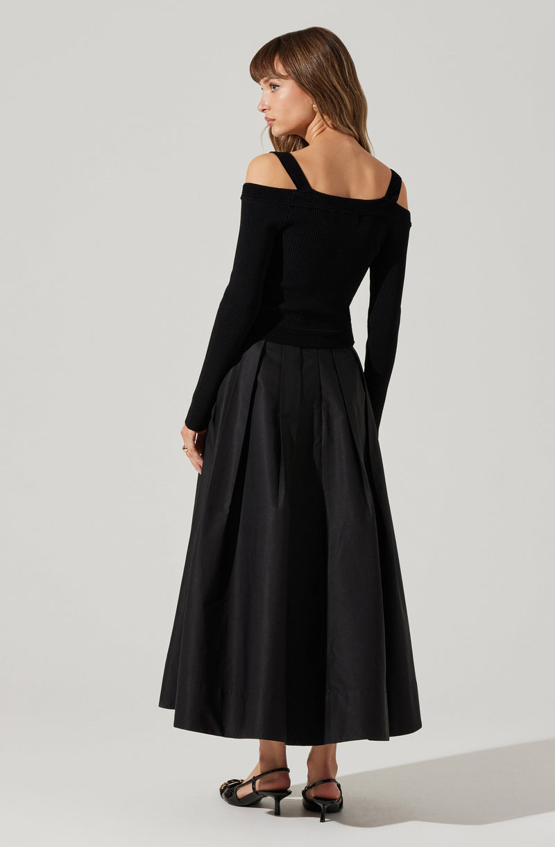 A woman is wearing a black off-the-shoulder sweater with shoulder straps, paired with a long, high-waisted, full-length black skirt with pleats. She is also wearing black sandals with a small heel.