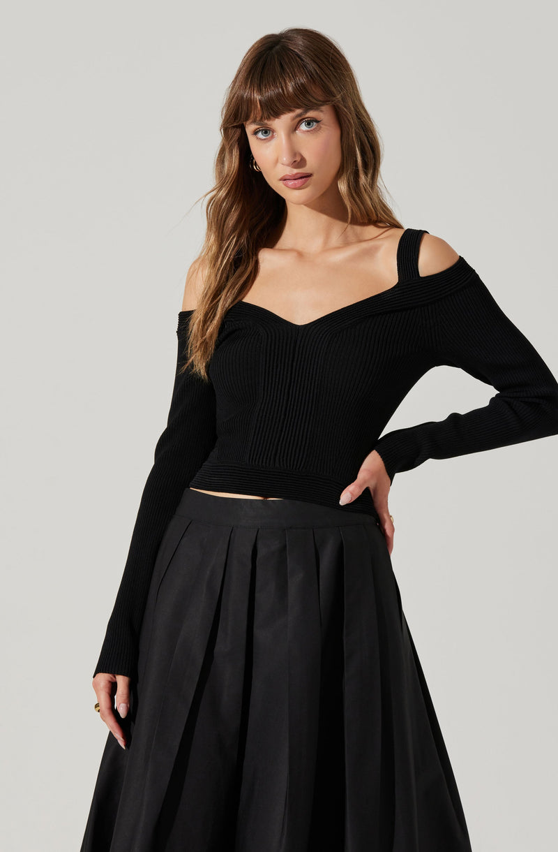 A young woman is wearing a black, fitted, long-sleeve sweater with a V-neck and off-the-shoulder design. The sweater has a ribbed texture and one strap over each shoulder. She pairs it with a high-waisted, black pleated skirt that falls below the knees.