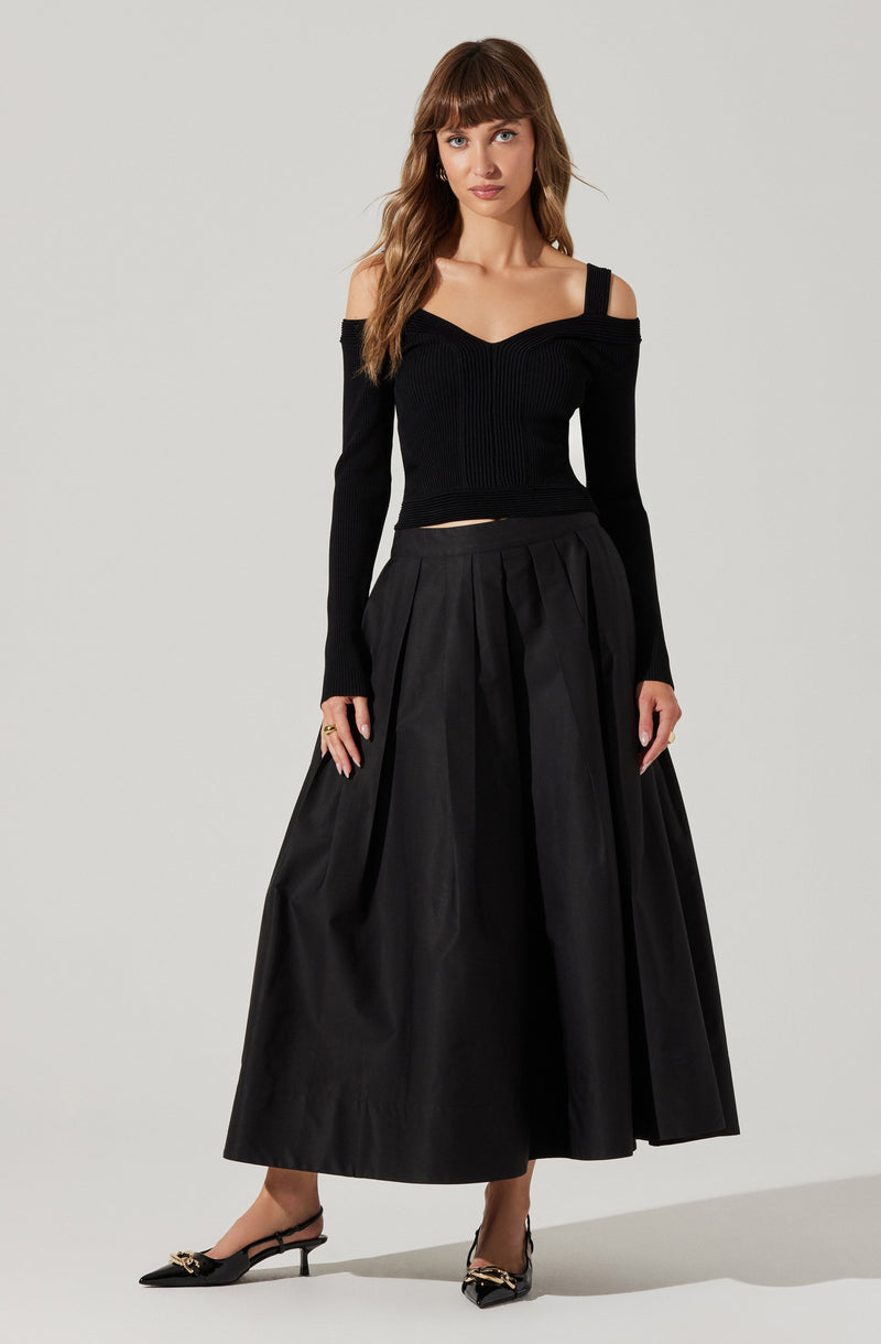 A woman is wearing a fitted, long-sleeved black sweater with a V-neckline and shoulder cut-outs. She pairs it with a high-waisted, full-length black skirt with pleats. She completes the outfit with black pointed-toe heels featuring a decorative chain detail across the front.