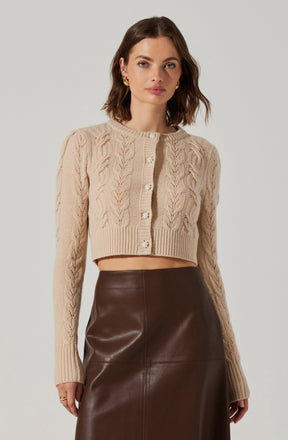 A woman is wearing a beige, long-sleeved cable-knit cardigan with a round neckline. The cardigan features an intricate pattern of cables and is decorated with shiny, pearl-like buttons down the front. She is also wearing a high-waisted, dark brown leather skirt that reaches below her knees.