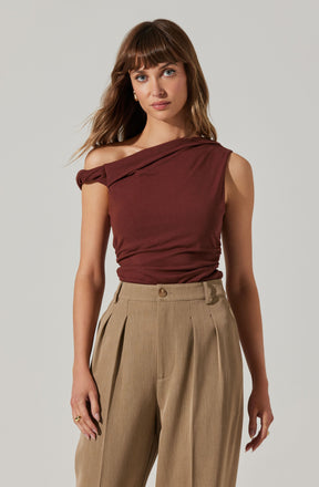 A woman is wearing a sleeveless, asymmetrical burgundy top that drapes over one shoulder, creating an off-shoulder look on one side. She is also dressed in high-waisted, beige pleated trousers.