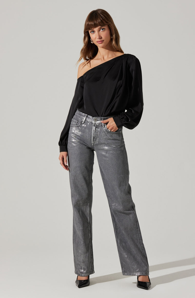 A woman is wearing a black, one-shoulder, long-sleeve blouse with a silky texture. She pairs it with high-waisted, straight-leg gray jeans that have a metallic sheen. She completes the look with black pointed-toe heels.