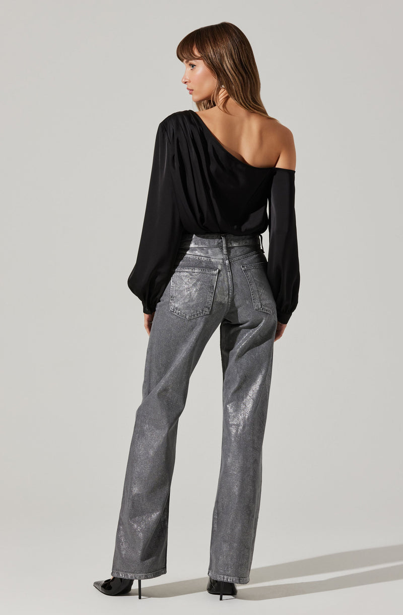 A woman is wearing a black off-the-shoulder top with long, slightly puffed sleeves that gather at the wrists. The top has a draped, asymmetrical neckline that exposes her left shoulder and part of her back. She pairs it with high-waisted, metallic silver jeans with a slightly loose fit and back pockets. She is also wearing black high-heeled shoes.