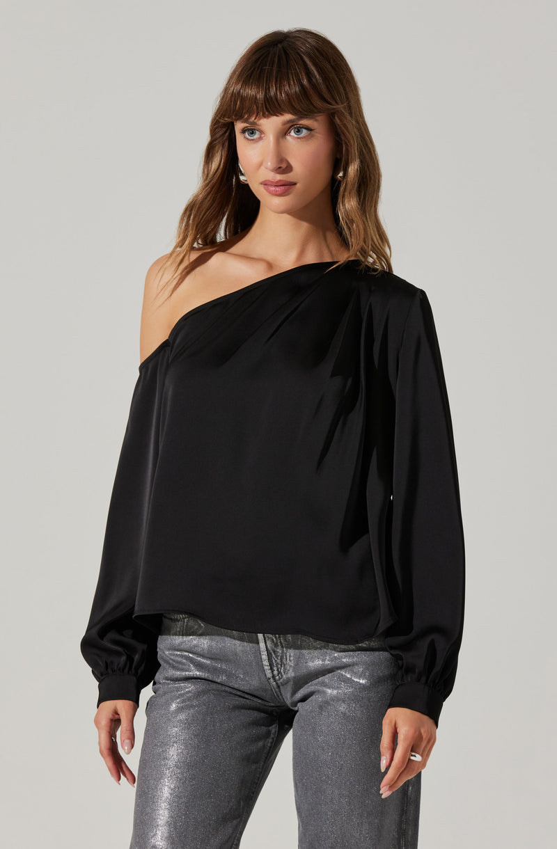 A woman is wearing a black, one-shoulder blouse with long, slightly puffed sleeves that are gathered at the wrists. The blouse features soft pleats and drapes elegantly across one shoulder, creating an asymmetrical neckline. She pairs it with metallic silver jeans.
