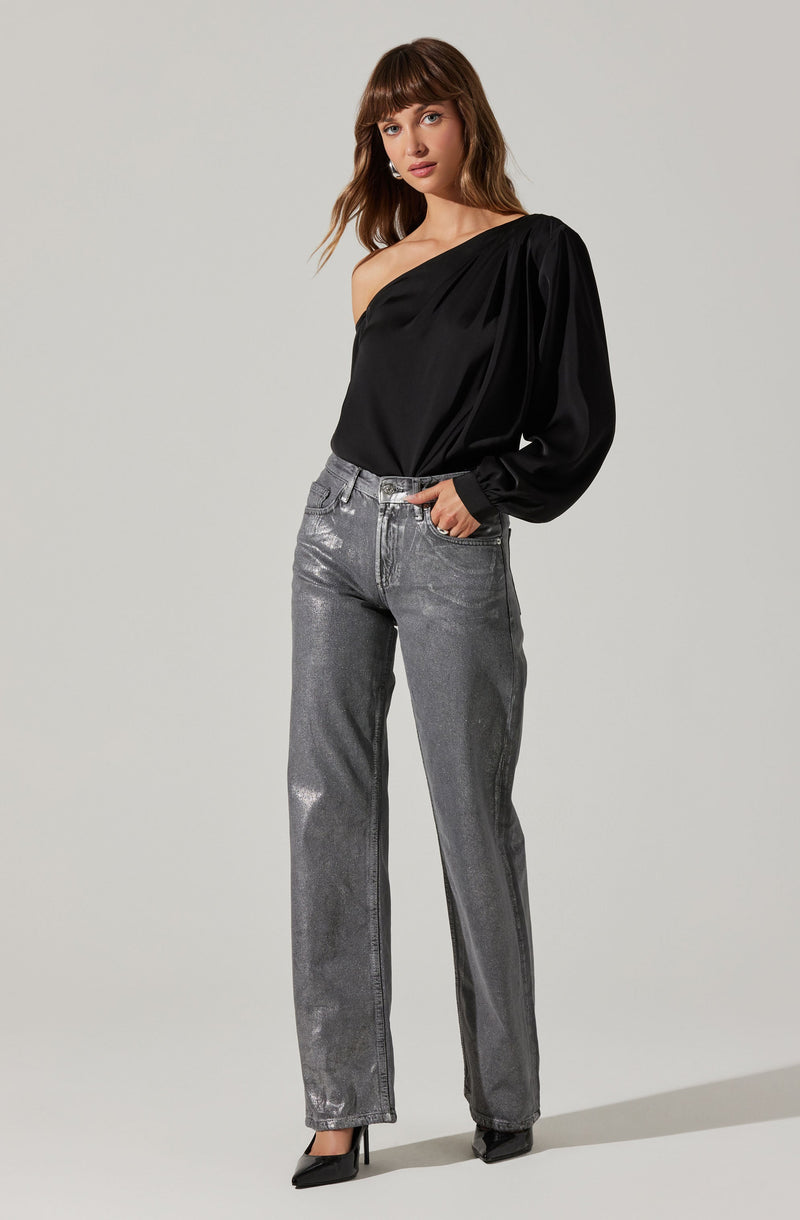 A woman is wearing a black, one-shoulder satin blouse with long, billowy sleeves. She pairs it with high-waisted, silver metallic jeans that have a straight-leg fit. She completes her look with black pointed-toe heels.