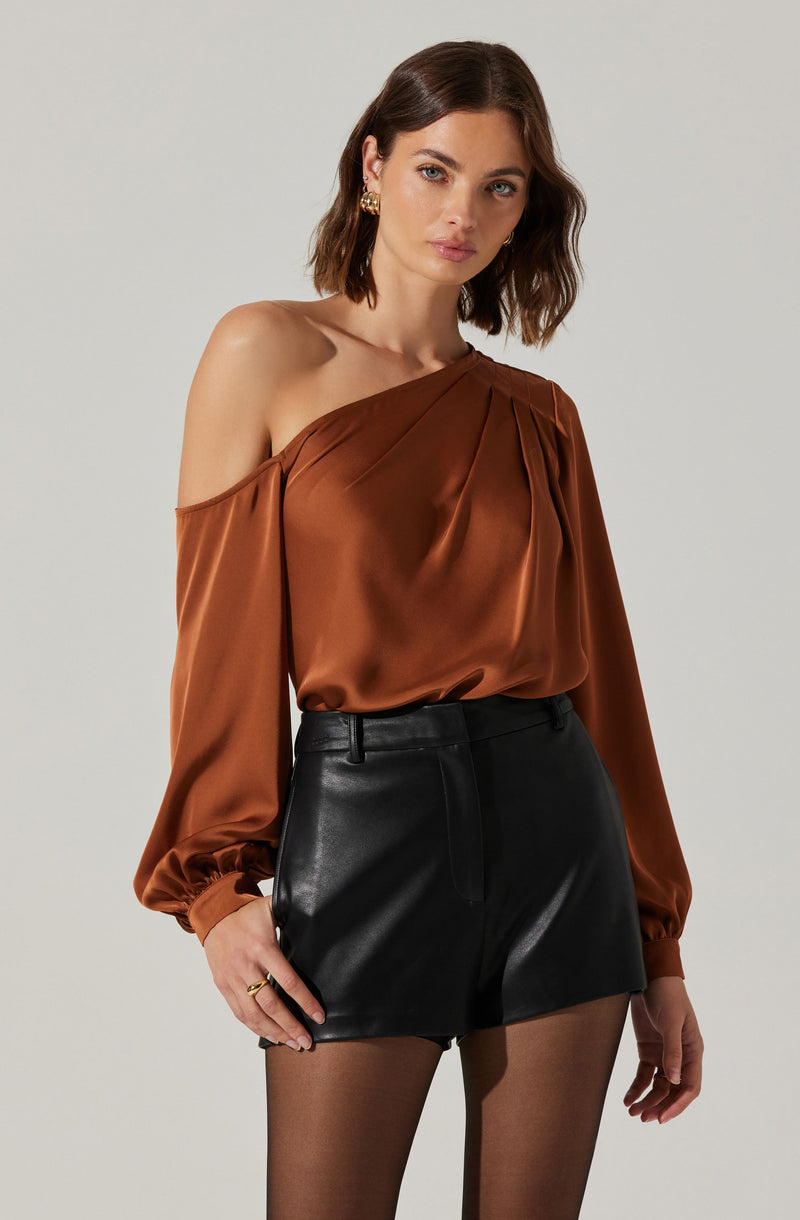 A woman is wearing an off-the-shoulder, long-sleeved, silky brown top with pleats at the shoulder and elasticized cuffs, paired with high-waisted black leather shorts.