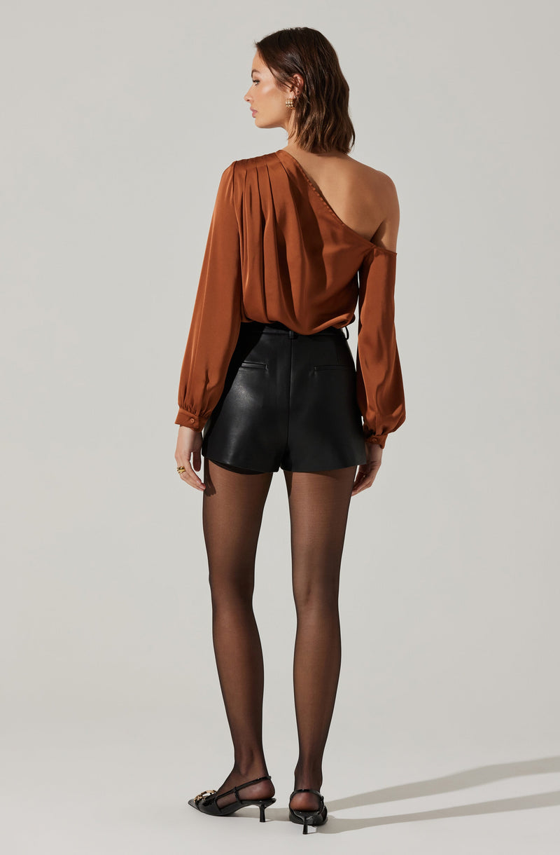 A woman is wearing a shiny, satin-like, off-the-shoulder, long-sleeve top in a copper brown color. She pairs it with high-waisted black leather shorts, sheer black stockings, and black high-heeled shoes with ankle straps.