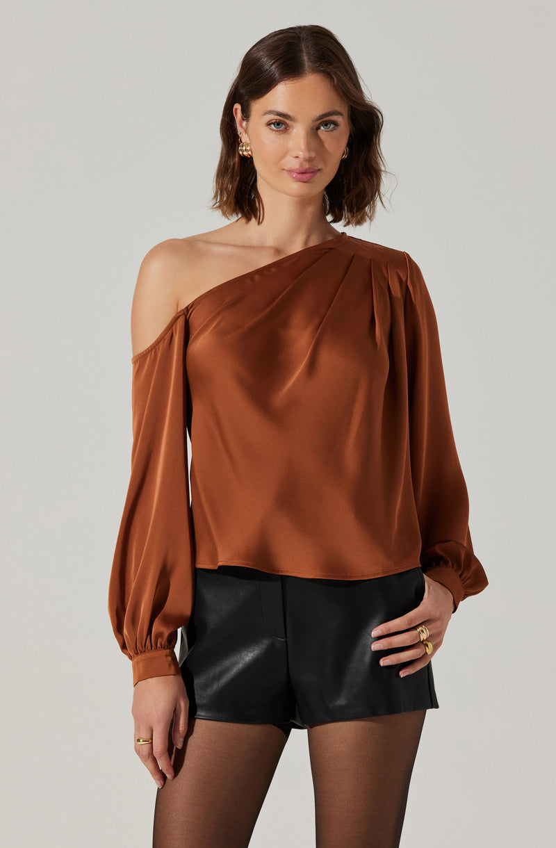 A woman is wearing a one-shoulder, rust-colored satin blouse with long, billowy sleeves gathered at the wrists. She pairs the top with high-waisted black faux leather shorts and sheer black tights. She accessorizes with gold hoop earrings and multiple rings.