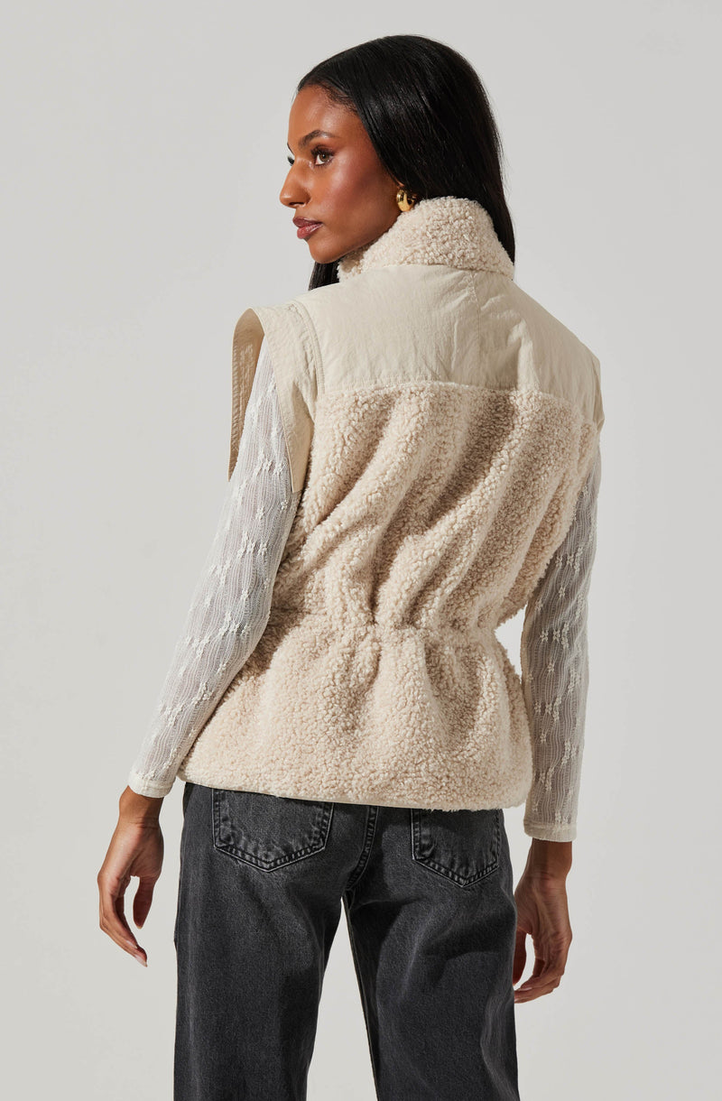 A woman is wearing a sleeveless, cream-colored fleece vest with a high collar. Underneath, she has on a long-sleeved, textured white top with a subtle pattern. She is also wearing dark gray, high-waisted jeans.