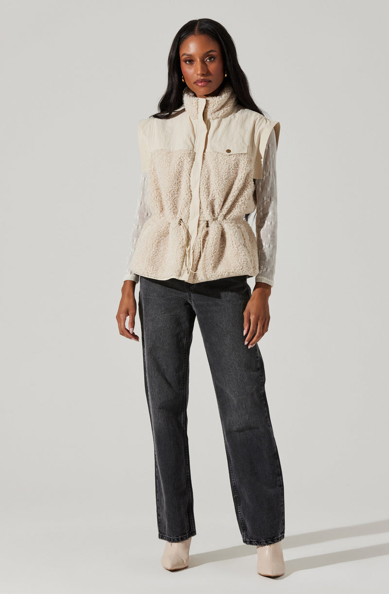 A woman is wearing a sleeveless, cream-colored vest with a high collar, featuring a zip closure in the front and a small pocket on the left chest with a button flap. She is also dressed in dark, loose-fitting jeans and light-colored ankle boots.