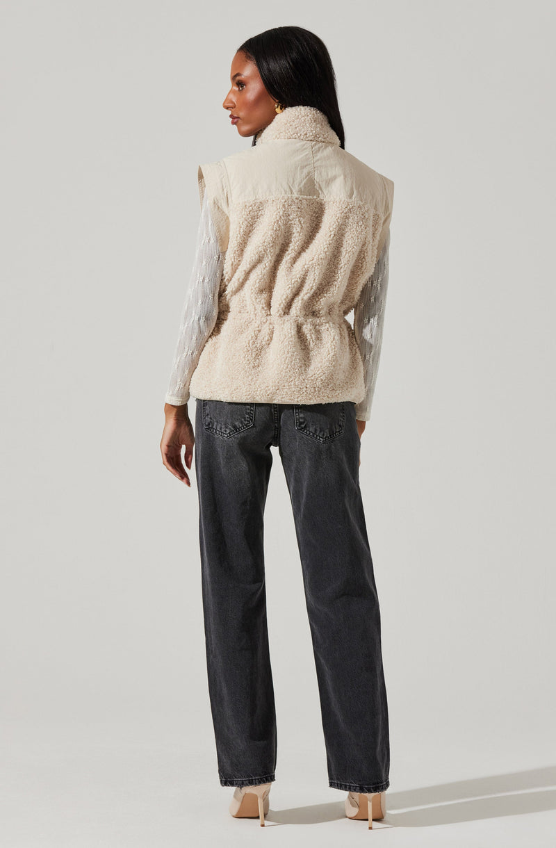 A woman is wearing a sleeveless, cream-colored vest with a fluffy, textured fabric. Underneath, she has on a long-sleeved, white blouse with subtle vertical stripes. She pairs these with high-waisted, dark gray jeans and beige high-heeled shoes.