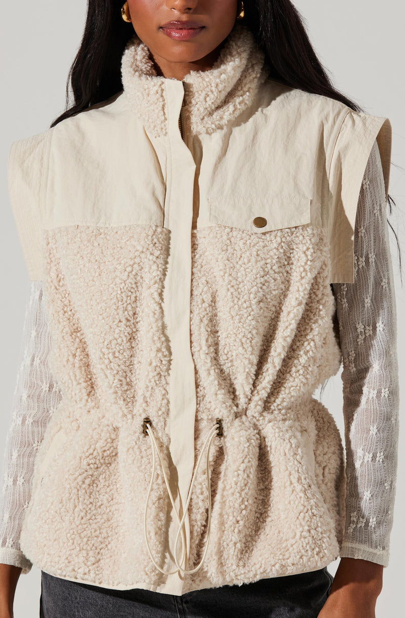 A woman is wearing a sleeveless, cream-colored vest made of a fluffy, sherpa-like material. The vest features a high collar, a front zipper, and a small chest pocket with a button flap. The vest also has drawstrings at the waist for adjustment. Underneath, she has on a long-sleeved, semi-sheer lace top with a floral pattern.