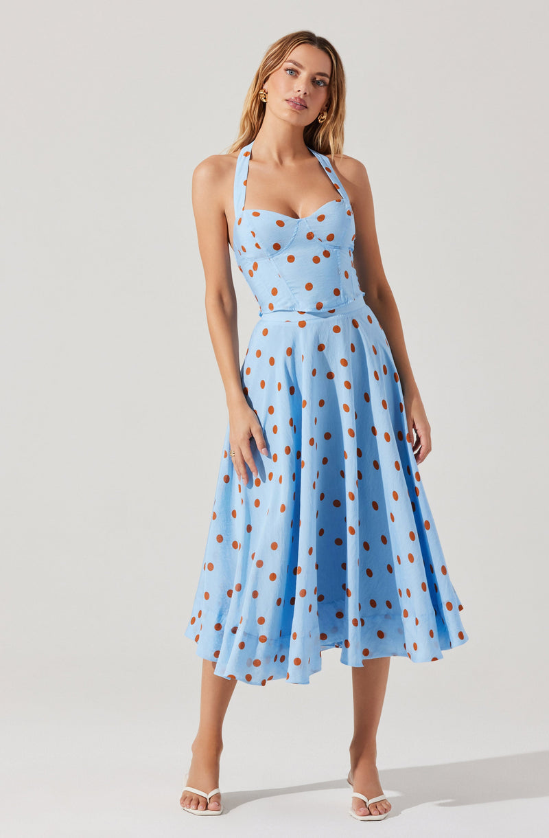 A woman is wearing a light blue dress with brown polka dots. The dress has a fitted bodice with a sweetheart neckline and a halter strap around the neck. The skirt is full and midi-length, with a pleated design that falls below the knees.