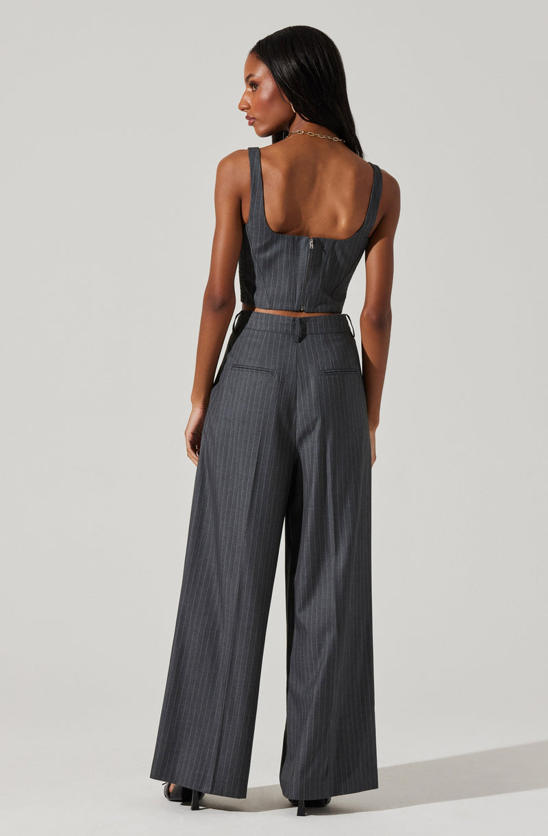 A woman is wearing a matching dark gray pinstripe sleeveless top with wide straps and high-waisted, wide-leg trousers. The top has a zipper down the back, and the trousers feature belt loops and two back pockets.