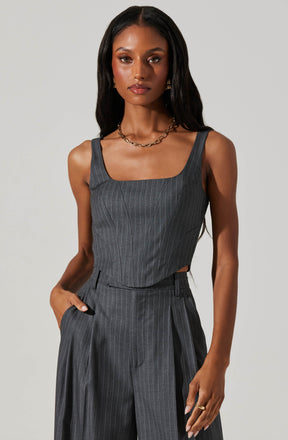 A woman is wearing a sleeveless, dark gray pinstriped top with a square neckline, paired with matching high-waisted, wide-leg pinstriped trousers. The outfit features subtle vertical pinstripes and side slits on the top. She accessorizes with a gold chain necklace, gold hoop earrings, and a gold ring on her right hand. Her long, dark hair is styled in loose waves.