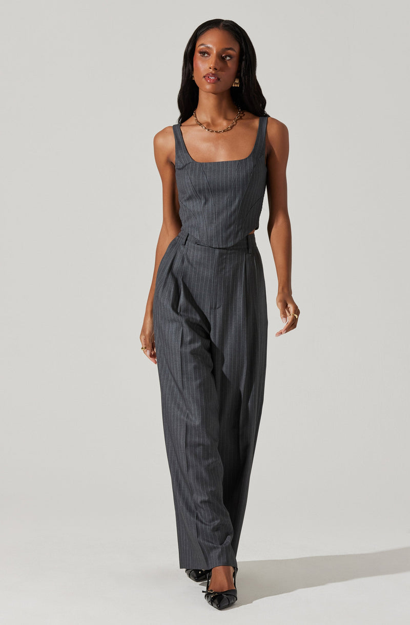 A woman is wearing a gray pinstripe sleeveless top with wide straps and matching high-waisted, loose-fitting pinstripe trousers. She has accessorized with gold hoop earrings, a gold chain necklace, and black pointed-toe shoes with a bow detail.