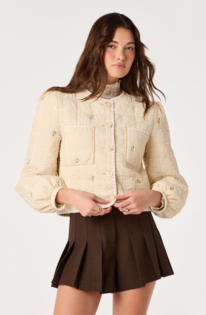 Woman in a cream textured jacket with floral embroidery and brown pleated shorts.