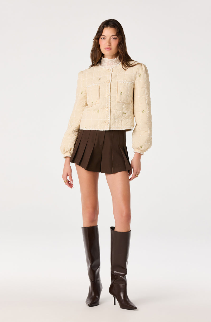 Cream embroidered textured jacket with puffed sleeves, brown pleated mini skirt, and tall brown boots.