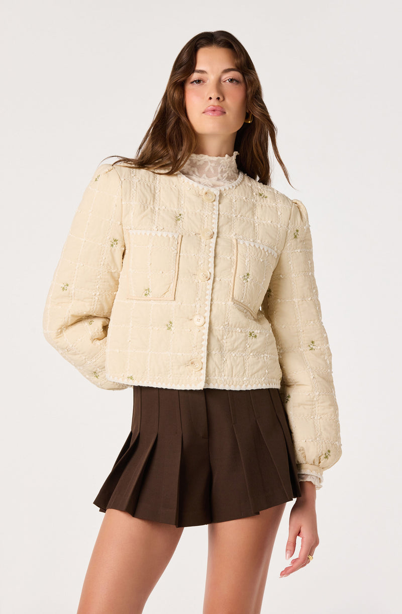 Woman in cream textured jacket with pockets and pleated brown shorts.