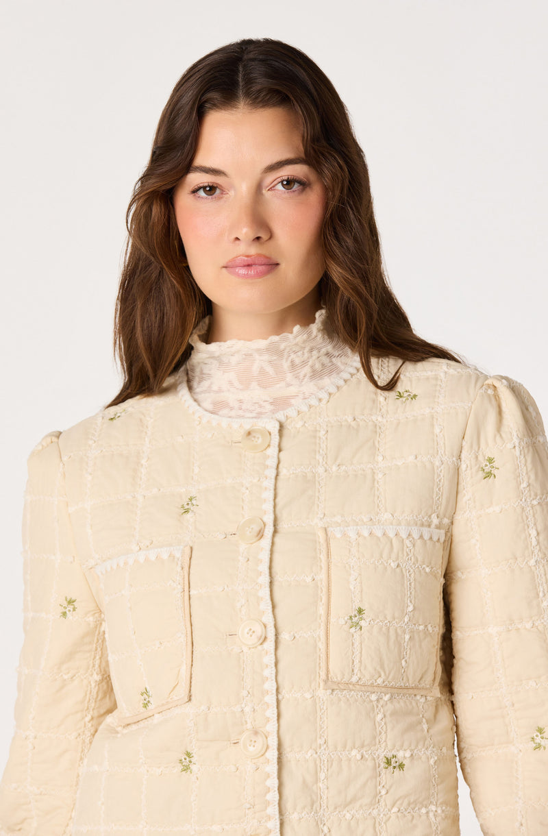 Cream-colored jacket with textured fabric, embroidered green floral details, and large front pockets.