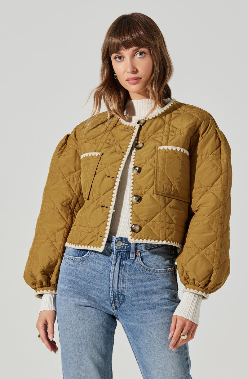 A woman is wearing a mustard yellow, quilted, cropped jacket with large buttons down the front and decorative white scalloped trim along the collar, pockets, and hem. Underneath, she has on a white ribbed sweater with ribbing at the cuffs. She pairs this with high-waisted, light-wash jeans.