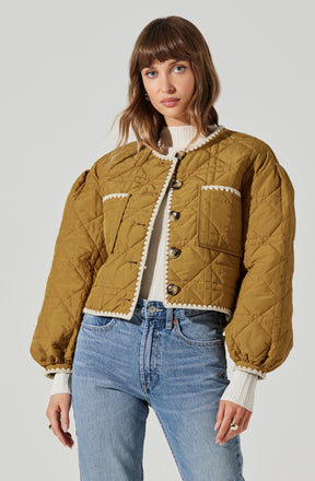A woman is wearing a mustard yellow, quilted, cropped jacket with large buttons down the front and decorative white scalloped trim along the collar, pockets, and hem. Underneath, she has on a white ribbed sweater with ribbing at the cuffs. She pairs this with high-waisted, light-wash jeans.