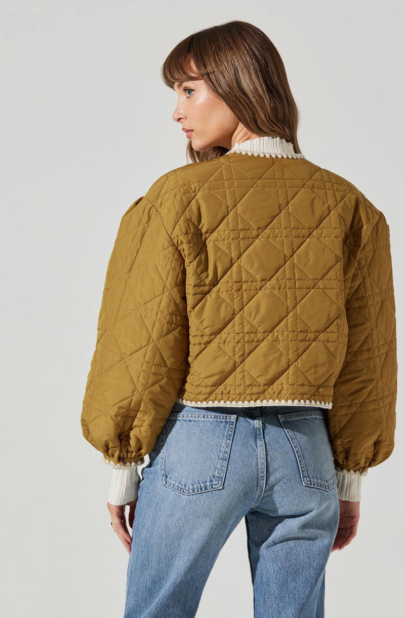 A woman is wearing a quilted, olive-green jacket with a slightly cropped fit. The jacket features puffy, long sleeves with elasticized cuffs, and a diamond-shaped quilted pattern throughout. She is also wearing a white, ribbed sweater underneath, with the collar and cuffs visible.