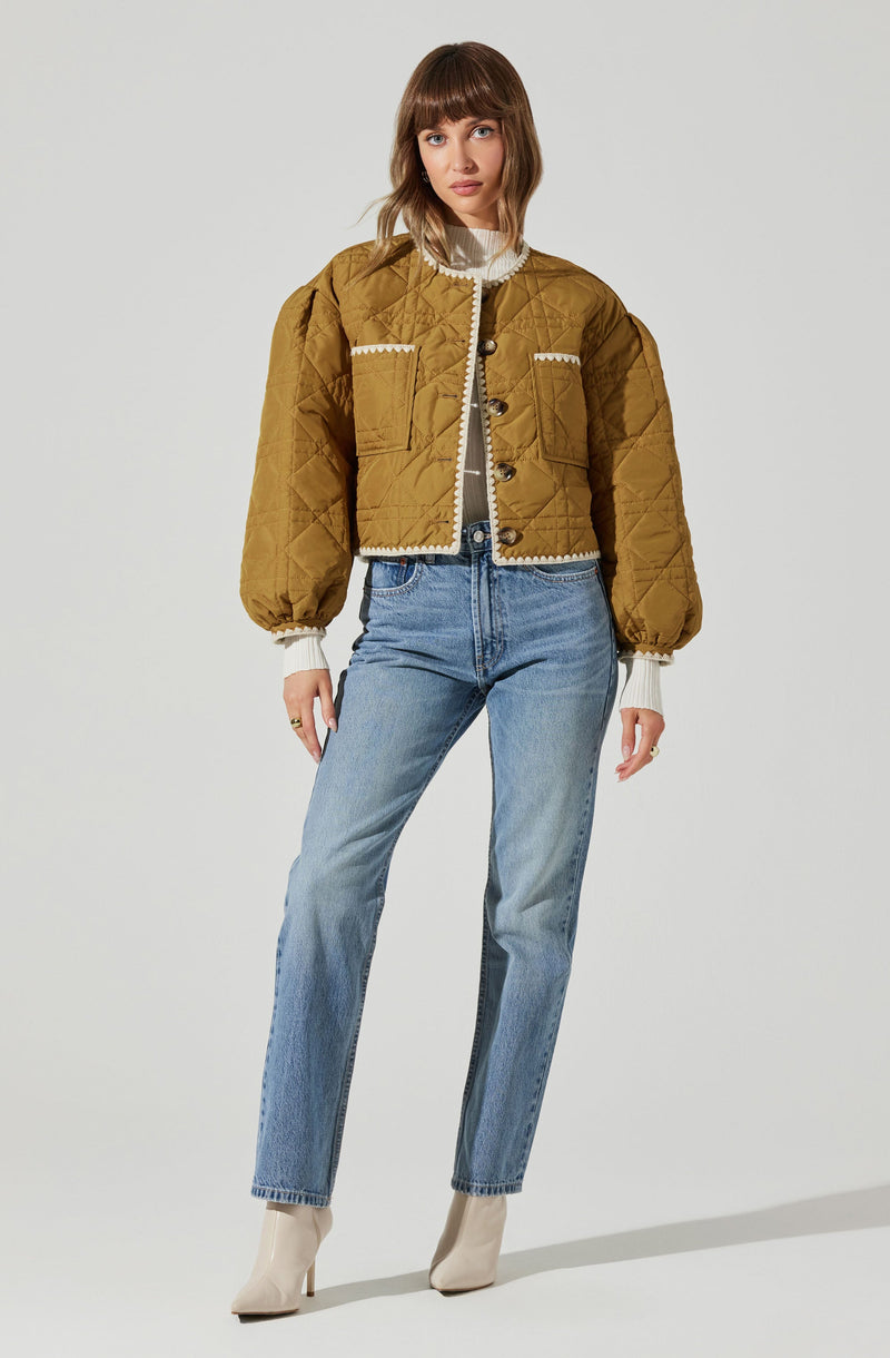 A woman is wearing a mustard-colored, quilted, cropped jacket with puffed sleeves, button closure, and decorative white piping details on the pockets and edges. She has on a white turtleneck sweater underneath, paired with high-waisted light blue jeans and beige ankle boots.
