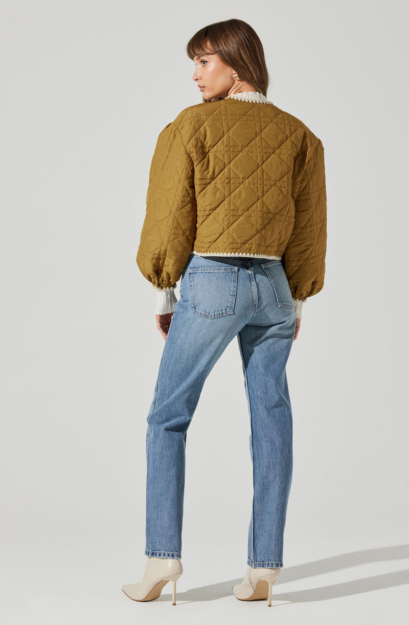 A woman is wearing a quilted, olive-green jacket with puffed sleeves and elastic cuffs, paired with light blue high-waisted jeans and beige high-heeled ankle boots. The jacket features a diamond quilt pattern and is cropped at the waist. She is also wearing a white, ruffled shirt underneath.