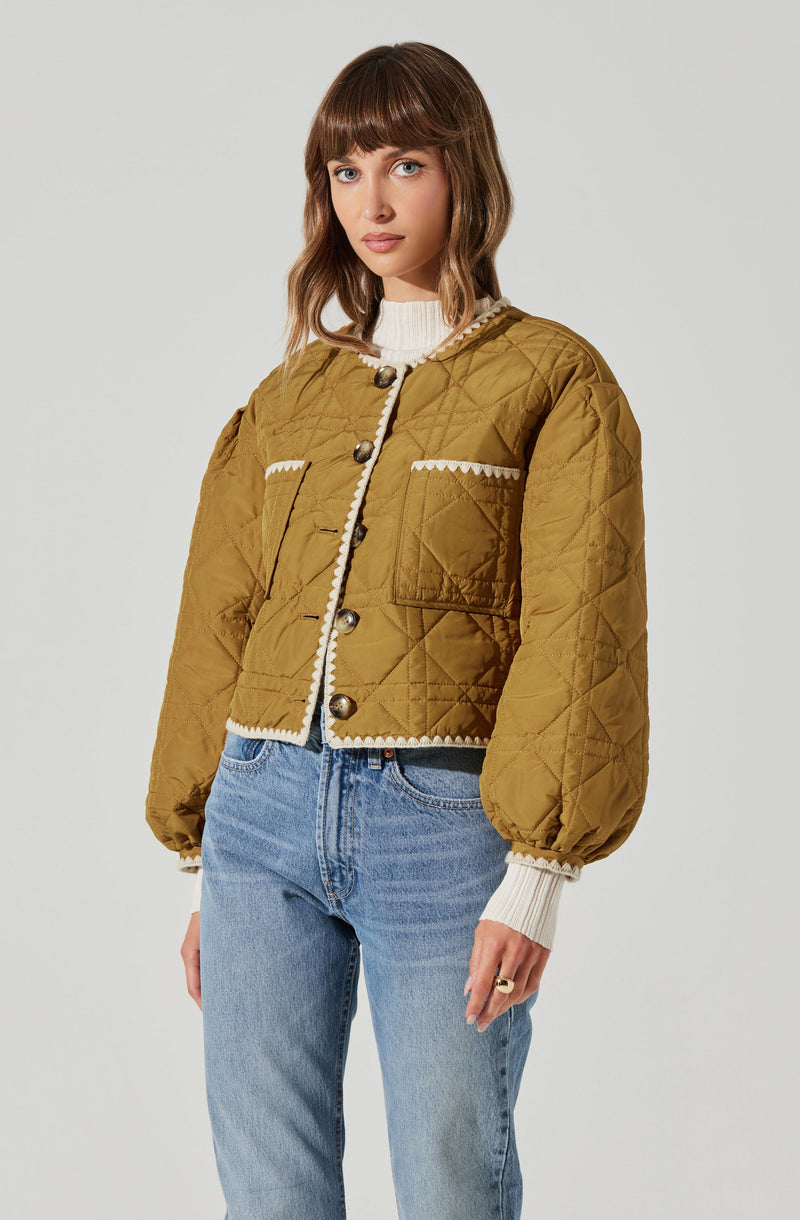 A woman is wearing a cropped, mustard yellow quilted jacket with puffed sleeves and large decorative buttons. The jacket has white decorative trim along the edges and pockets. Underneath, she has on a white ribbed turtleneck sweater with long sleeves. She is also wearing high-waisted, light-wash jeans.