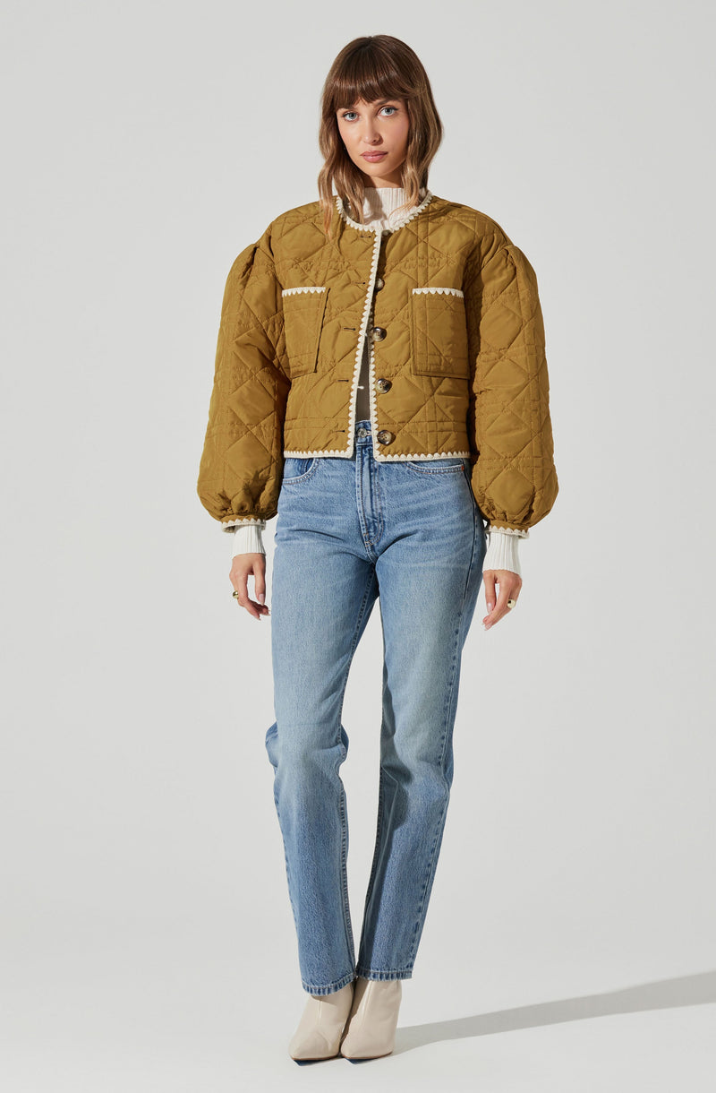 A woman is wearing a mustard yellow quilted jacket with puffed sleeves and decorative white trim along the collar, front pockets, and cuffs. She pairs the jacket with a white ribbed sweater, light-wash high-waisted jeans, and beige ankle boots.