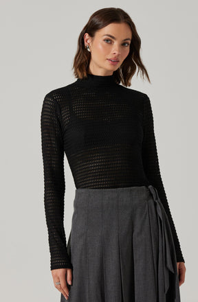 A woman is wearing a black, long-sleeved, high-neck sweater with a perforated, open-knit pattern. She pairs it with a high-waisted gray skirt that features vertical pleats and a matching fabric tie belt.