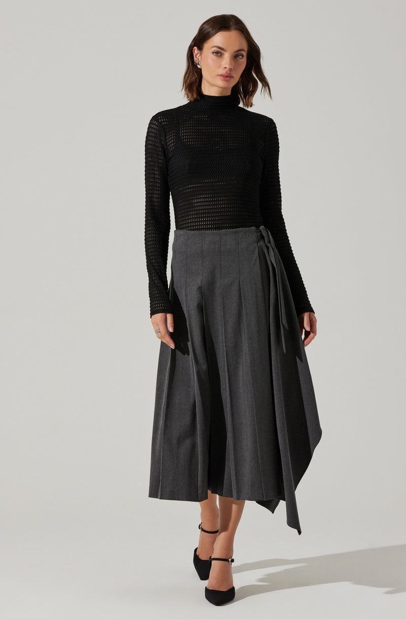 A woman is wearing a fitted black long-sleeve top with a high neckline, made of a see-through, open-knit fabric. She pairs it with a high-waisted, wide-leg gray skirt that features vertical pleats and a tied sash detail on one side. She completes her outfit with black ankle-strap high heels.