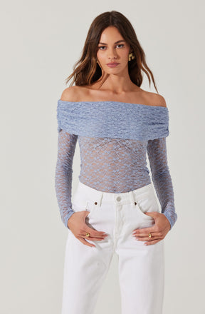 A woman is wearing an off-the-shoulder, light blue lace top with a wide, textured band around the shoulders. The top features a delicate, floral lace pattern throughout with fitted long sleeves. She pairs the top with high-waisted white jeans.
