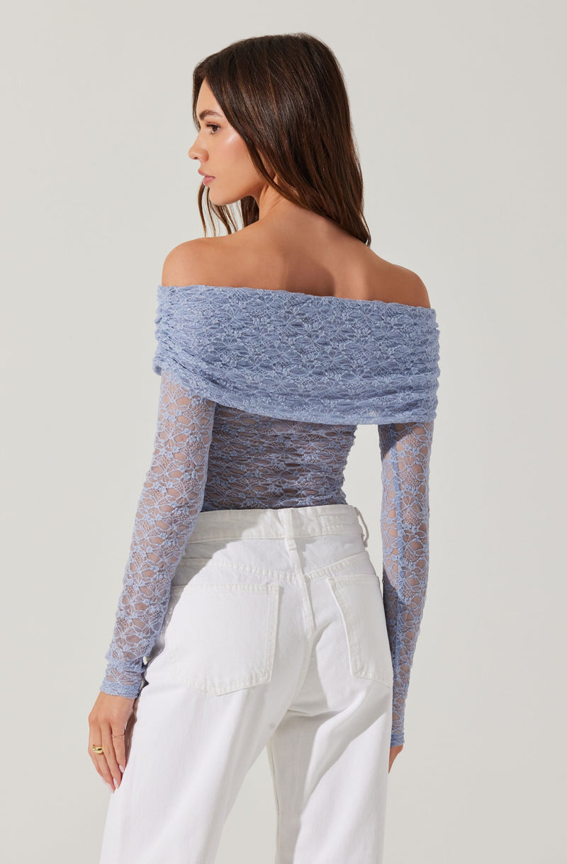 A woman is wearing an off-the-shoulder, long-sleeved lace top in a light blue color. The top features a wide, textured lace band around the shoulders and a fitted, sheer lace body with a floral pattern. She pairs it with high-waisted white pants.