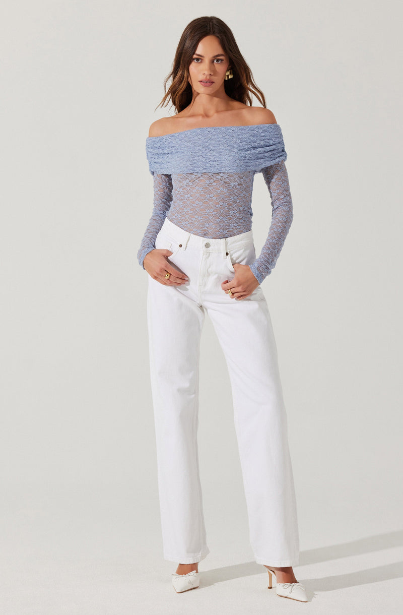 The woman is wearing an off-the-shoulder, long-sleeved top made of a textured, light blue fabric that is slightly see-through with a floral lace pattern. She is also wearing high-waisted, straight-leg white jeans with a button and zipper closure. She has on white heels with a pointed toe and a bow detail on the front.