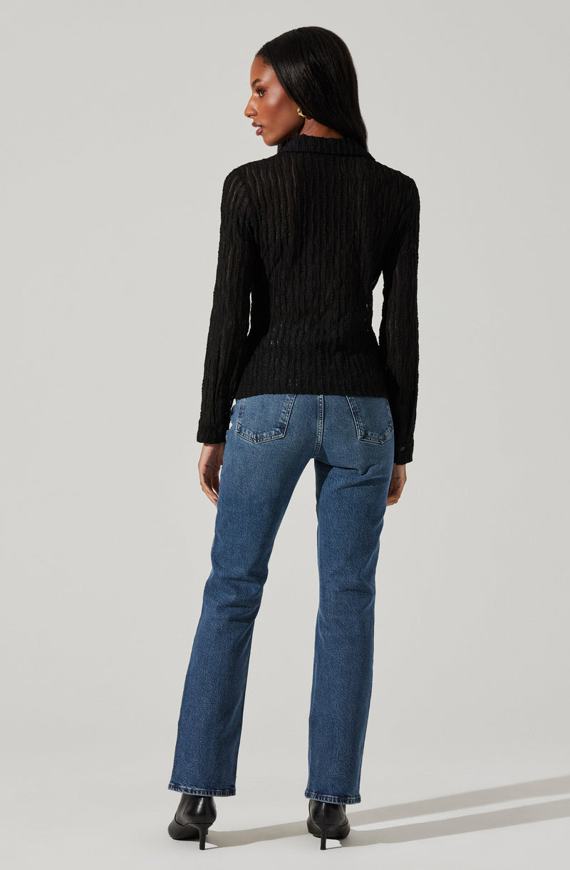 A woman is wearing a black, long-sleeved, textured sweater with a high neckline, paired with high-waisted blue jeans that have visible stitching and back pockets. She is also wearing black heeled ankle boots.