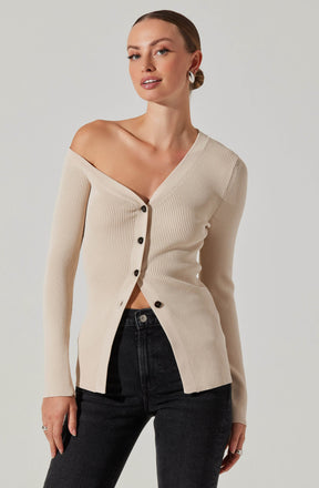 A woman is wearing a beige, ribbed, V-neck button-up cardigan with long sleeves. The cardigan is slightly off-the-shoulder on her left side, and the buttons are dark-colored. She pairs the cardigan with high-waisted black jeans and has her hair styled in a sleek bun. She is also wearing large hoop earrings.