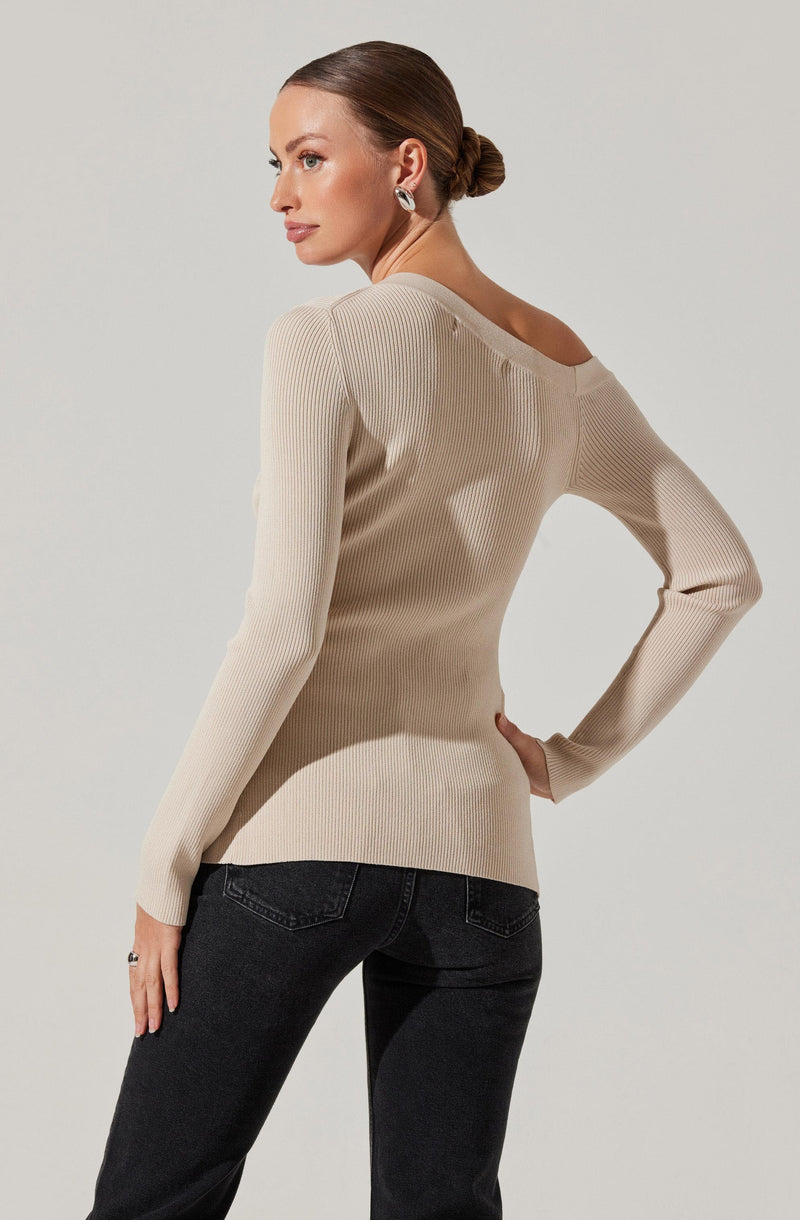 A woman is wearing a long-sleeved, beige ribbed knit sweater with a wide, rounded neckline. She has paired it with black jeans.