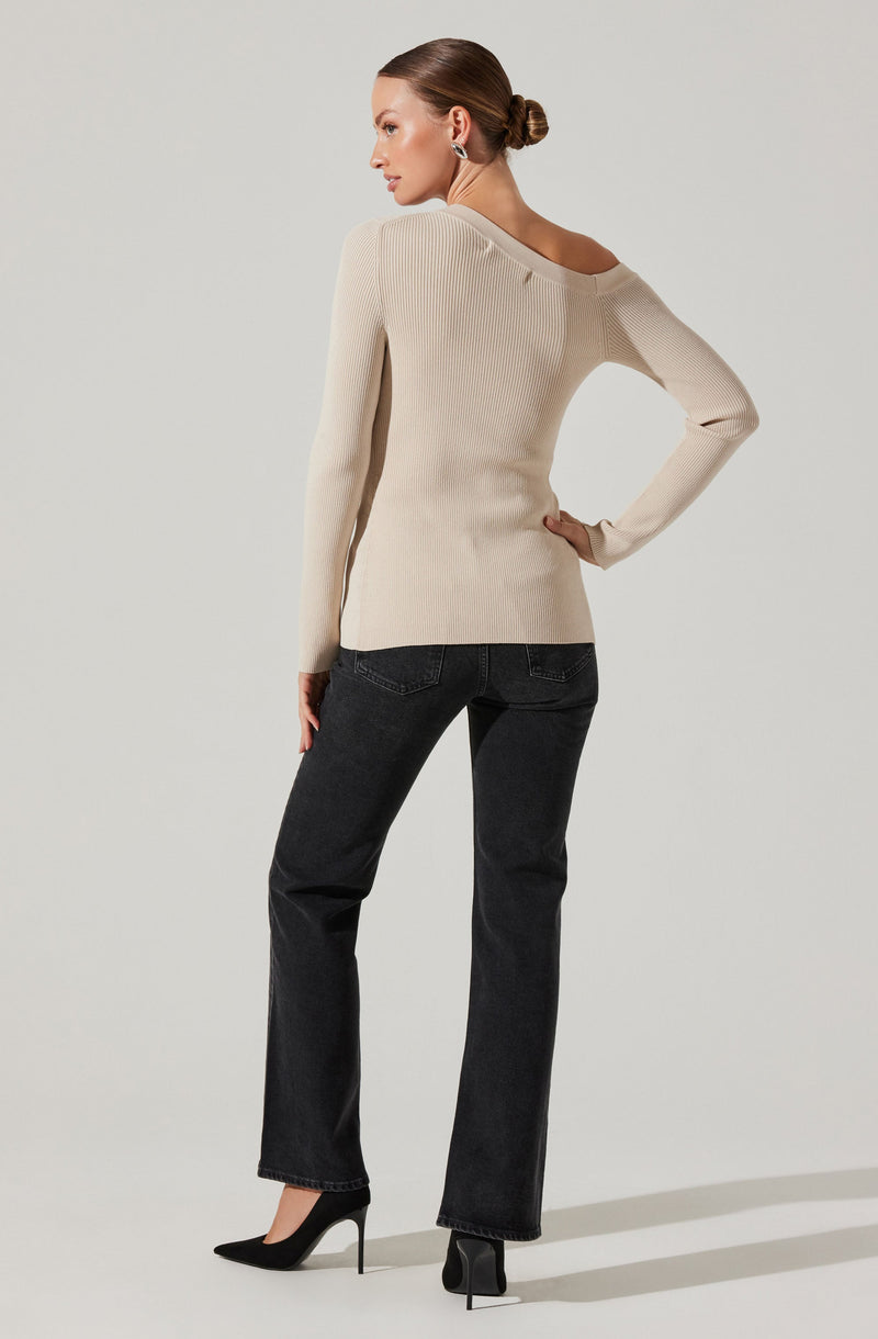 A woman is wearing a cream-colored, ribbed knit sweater with an asymmetrical neckline that falls off one shoulder. She pairs it with dark high-waisted jeans and black high-heeled shoes. Her hair is styled in a low bun, and she has on large earrings.
