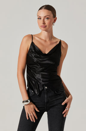 A woman is wearing a black satin sleeveless top with thin spaghetti straps and a draped neckline. She pairs it with high-waisted black jeans. She has accessorized with a large silver cuff bracelet on her right wrist, a ring on her right hand, and hoop earrings.