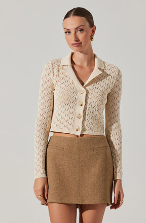 A woman wearing a cream-colored, long-sleeved, button-up cardigan with a diamond-shaped open-knit pattern. The cardigan has a collar and is cropped, ending just above her waist. She pairs it with a high-waisted, brown, woolen mini skirt that has a simple, straight cut and two welt pockets at the front.