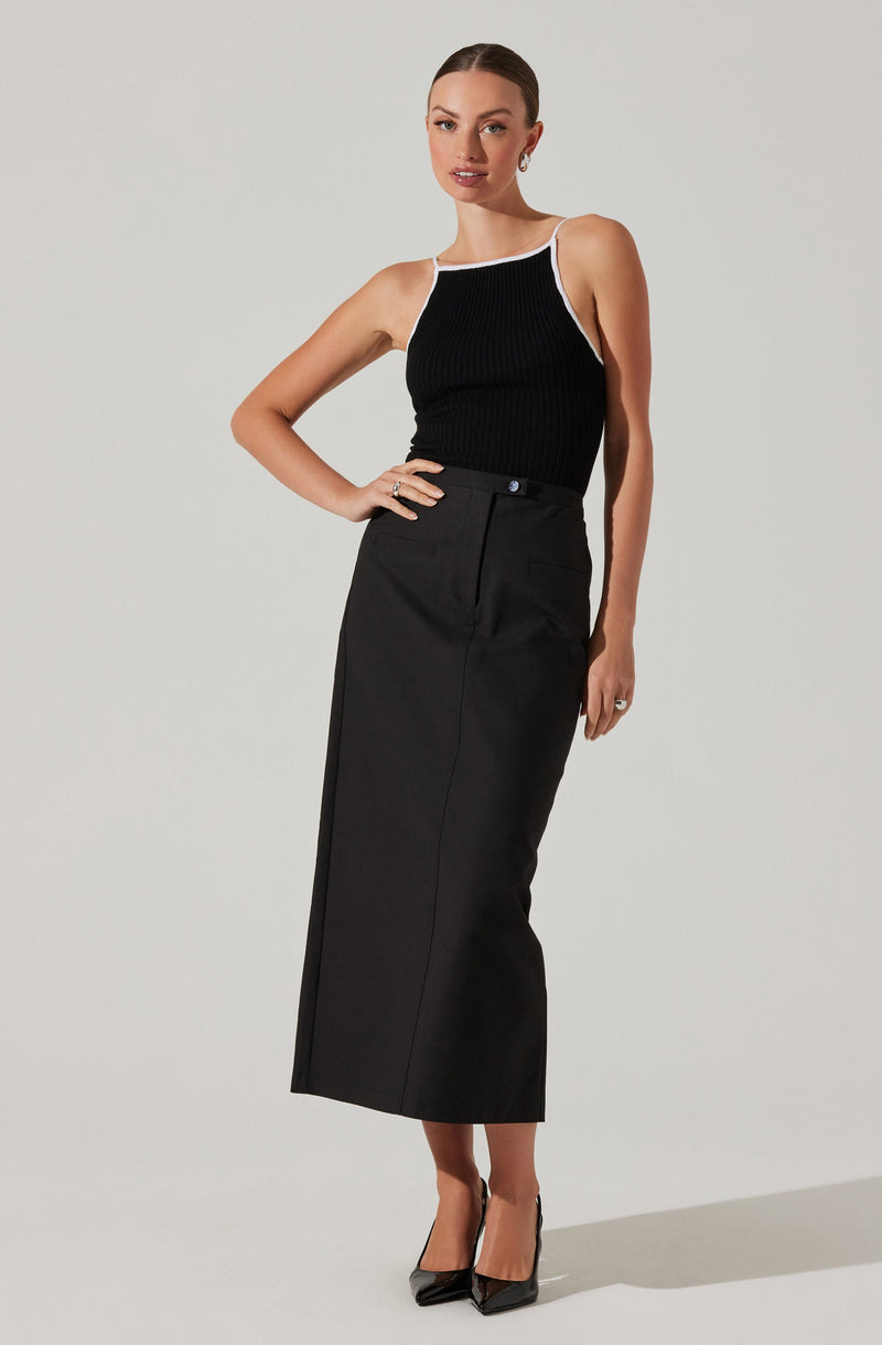 A woman is wearing a sleeveless black ribbed top with white trim along the neckline and armholes. She pairs it with a high-waisted, long black skirt that features a front seam, side slit, and a small button at the waist. She completes the outfit with black pointed-toe high heels.