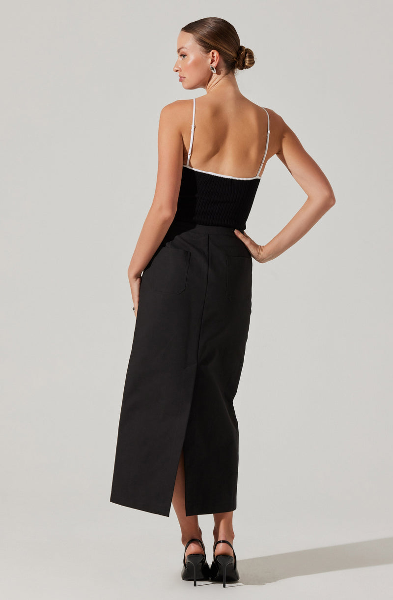 A woman is wearing a black sleeveless top with thin white straps and a white trim along the top edge. She pairs it with a high-waisted black skirt that has a front slit and side pockets. She completes her outfit with black high-heeled shoes.